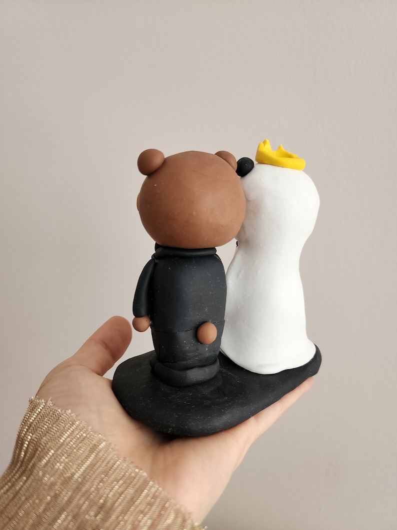 May include: A brown bear wearing a black suit and a white polar bear wearing a white dress and a yellow crown. The bears are standing on a black base.