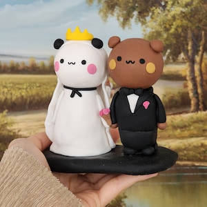 May include: A white panda bride with a yellow crown and a black bow tie, and a brown bear groom with a black bow tie and a pink flower, stand on a black cake base. The figures are made of clay and are a wedding cake topper.