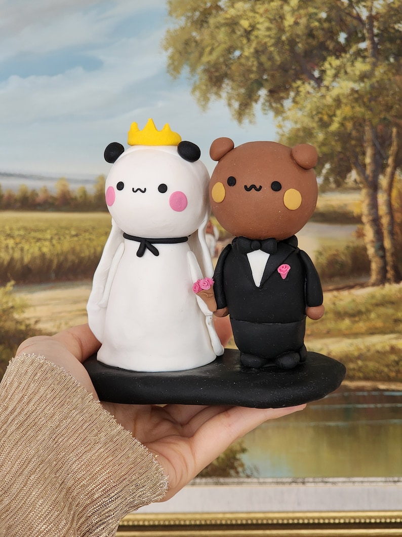 May include: A white panda bride with a yellow crown and a black bow tie, and a brown bear groom with a black bow tie and a pink flower, stand on a black cake base. The figures are made of clay and are a wedding cake topper.