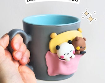 May include: A gray ceramic mug with a light blue interior. The mug has a 3D design of a panda and a brown bear in a pink blanket. The text "BUBU-DUDU ON A MUG" is on a white sticker.