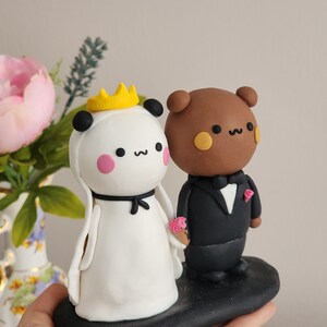 May include: A white panda bride with a yellow crown and a black bow, holding a pink flower bouquet, stands next to a brown bear groom wearing a black tuxedo and a pink flower boutonniere. The couple is standing on a black base.