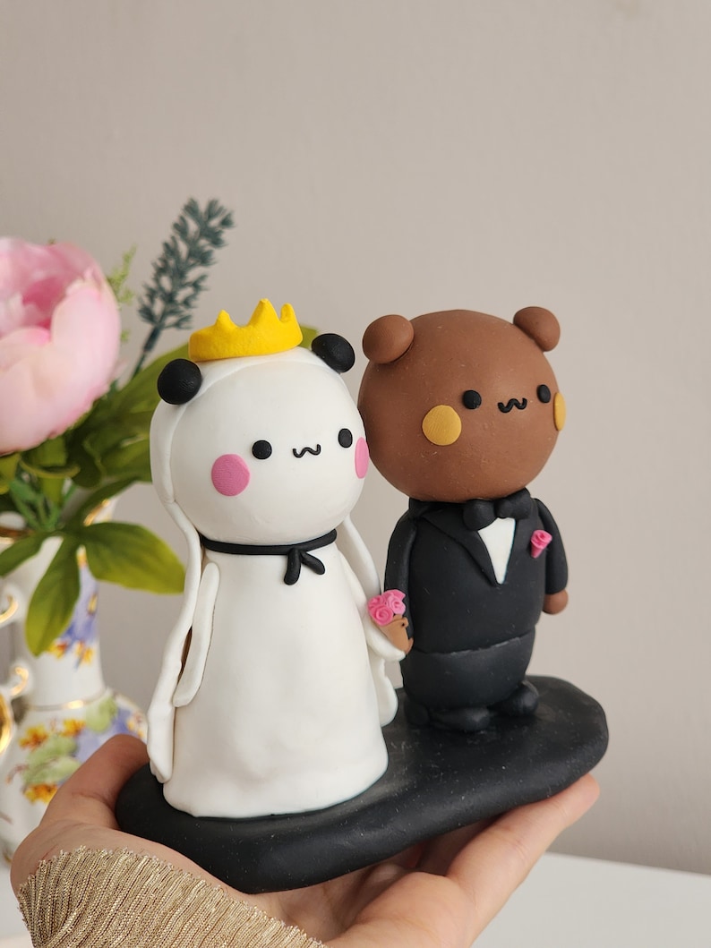 May include: A white panda bride with a yellow crown and a black bow, holding a pink flower bouquet, stands next to a brown bear groom wearing a black tuxedo and a pink flower boutonniere. The couple is standing on a black base.