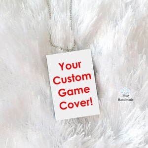 Puede incluir: Un colgante rectangular blanco con el texto "Your Custom Game Cover!" en letras rojas. El colgante está colgado de una cadena plateada y se encuentra sobre una superficie blanca y esponjosa. El texto "Blue Handmade" es visible en la esquina inferior derecha de la imagen.