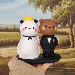 May include: A white panda and a brown bear wedding cake topper. The panda is wearing a white dress with a yellow crown and the bear is wearing a black tuxedo. The cake topper is on a black base.