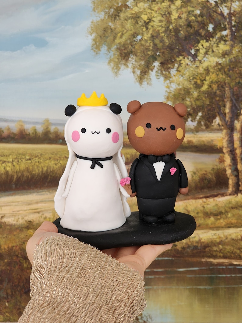 May include: A white panda and a brown bear wedding cake topper. The panda is wearing a white dress with a yellow crown and the bear is wearing a black tuxedo. The cake topper is on a black base.