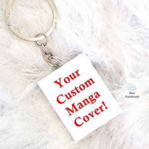 Könnte beinhalten: Weißer quadratischer Schlüsselanhänger mit rotem Text "Your Custom Manga Cover!" an einem silbernen Schlüsselring.