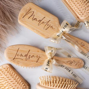May include: A collection of wooden hairbrushes, some personalized with names like "Madelyn" and "Lindsay." Each brush is tied with a sheer ribbon printed with "Just For You." The brushes vary in shape and size, with natural bristles.