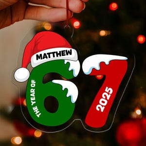 May include: A clear Christmas ornament with a red Santa hat and the name "MATTHEW" in white. The ornament features the numbers "67" in green and "2025" in red, with the text "THE YEAR OF" in green. The ornament is held in front of a Christmas tree.