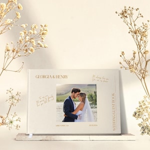 Puede incluir: Un libro de invitados de boda blanco con letras doradas. La portada presenta los nombres "Georgia & Henry" y una foto de una pareja en un campo. El libro tambi&eacute;n incluye la fecha "06.13.2021" y la ubicaci&oacute;n "Lake Tahoe, CA".