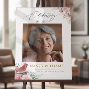 May include: A memorial sign on an easel, featuring a photo of a woman with gray hair. The sign reads "Celebrating the Life Of" and "Nancy Williams, August 18, 1946 - June 7, 2025." Floral and bird illustrations adorn the corners.