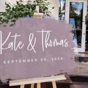Pu&ograve; includere: Un cartello nuziale trasparente su un cavalletto di legno. Il cartello presenta i nomi "Kate & Thomas" in corsivo bianco su un design a pennellate color malva. La data "SEPTEMBER 05, 2024" &egrave; stampata sotto. Sullo sfondo si vede del verde.