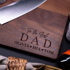 May include: A wooden cutting board with the text "To the Best Dad Olivia Mia Tom" engraved on it. The board is brown and has a natural wood grain. There is a knife and a bottle of wine in the background.