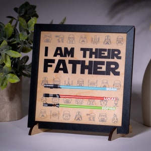 May include: A framed wooden sign with the text "I AM THEIR FATHER" and lightsaber illustrations. The sign includes names below the lightsabers. The frame is black, and the background is a light brown color.