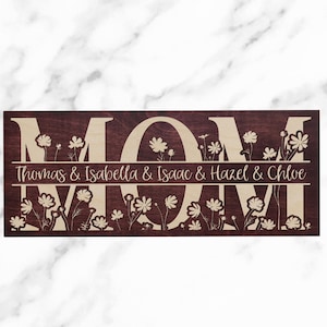 May include: A wooden sign with the word "MOM" in large, beige letters on a dark brown background. The letters are surrounded by a floral design. The text "Thomas & Isabella & Isaac & Hazel & Chloe" is written in beige letters below the word "MOM".