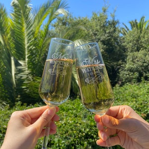 May include: Two champagne flutes with the words "Mr. Watkins 7.16.2021" and "Mrs. Watkins 7.16.2021" etched on them. The flutes are filled with champagne and are being held up in a toast.