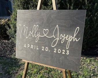 May include: A dark gray wooden sign with the names "Molly" and "Joseph" written in white cursive script, connected by a heart symbol. The date "APRIL 20, 2023" is written below the names.