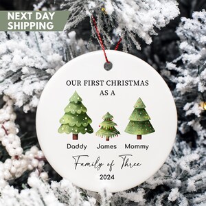 May include: White ceramic Christmas ornament with green, red, and white illustrated Christmas trees. The ornament reads "Our First Christmas as a Family of Three 2024".