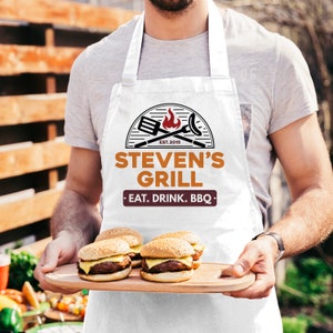 May include: White apron with a personalized logo that says "Steven's Grill - Eat. Drink. BBQ."  The logo features a grill, spatula, and flames. A person is holding a wooden tray with four cheeseburgers.