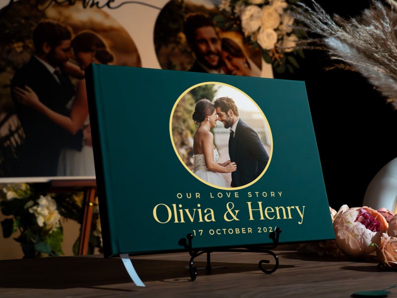May include: A dark green guest book with a gold circle containing a photo of a couple. The text "Our Love Story Olivia & Henry 17 October 2021" is printed in gold on the front cover.