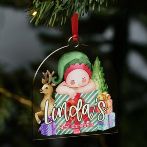 May include: A clear, arch-shaped acrylic Christmas ornament featuring a watercolor illustration of a baby in a green elf hat resting on a green and white striped gift box, a reindeer, and stacked presents with the text Linda's First Christmas 2024.