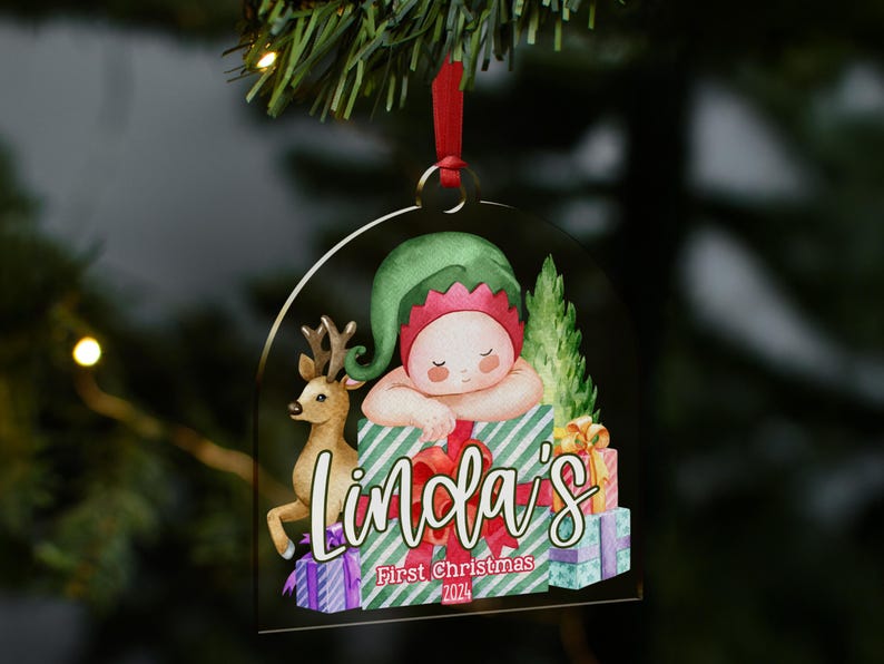 May include: A clear, arch-shaped acrylic Christmas ornament featuring a watercolor illustration of a baby in a green elf hat resting on a green and white striped gift box, a reindeer, and stacked presents with the text Linda's First Christmas 2024.