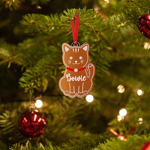 May include: A brown gingerbread cat ornament with a red bow and the name "Bowie" written on it. The ornament is hanging on a Christmas tree with red and white lights.