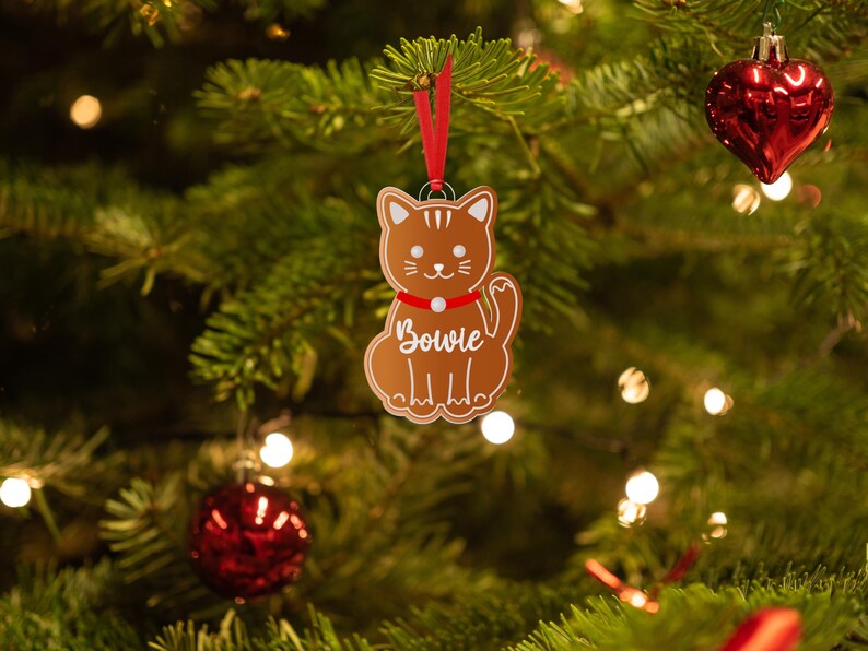 May include: A brown gingerbread cat ornament with a red bow and the name "Bowie" written on it. The ornament is hanging on a Christmas tree with red and white lights.