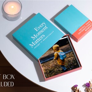 K&ouml;nnte beinhalten: Eine t&uuml;rkisfarbene Geschenkbox mit dem Text "Every Moment Matters Frame It Beautifully" auf dem Deckel. Die Box ist ge&ouml;ffnet und enth&auml;lt ein Fotoalbum mit einem roten Einband. Das Album ist auf einer Seite mit einem Foto eines Kindes in einem gelben Regenmantel und einem blauen Rucksack ge&ouml;ffnet. Die Box steht auf einem wei&szlig;en Tisch mit einer Kerze in einem Glasgef&auml;&szlig; im Hintergrund.