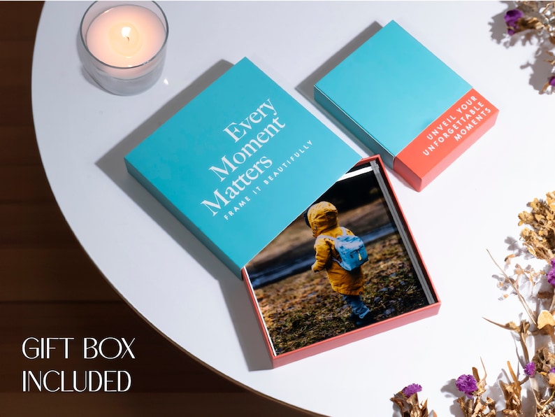 K&ouml;nnte beinhalten: Eine t&uuml;rkisfarbene Geschenkbox mit dem Text "Every Moment Matters Frame It Beautifully" auf dem Deckel. Die Box ist ge&ouml;ffnet und enth&auml;lt ein Fotoalbum mit einem roten Einband. Das Album ist auf einer Seite mit einem Foto eines Kindes in einem gelben Regenmantel und einem blauen Rucksack ge&ouml;ffnet. Die Box steht auf einem wei&szlig;en Tisch mit einer Kerze in einem Glasgef&auml;&szlig; im Hintergrund.