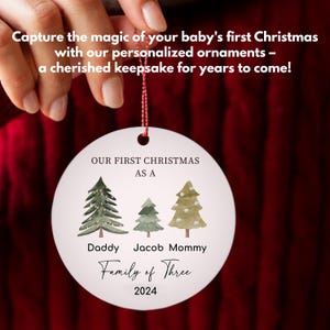May include: White round Christmas ornament with a watercolor design of three green Christmas trees. The ornament reads "Our First Christmas as a Family of Three 2024" with the names "Daddy", "Jacob", and "Mommy" under the trees.