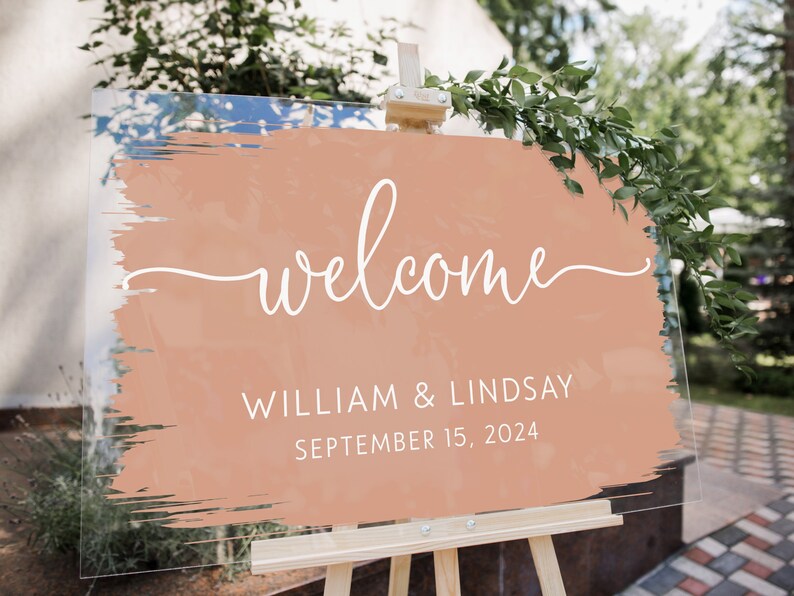 Pu&ograve; includere: Un cartello di benvenuto in acrilico trasparente con un design a pennellate color pesca. La parola "welcome" &egrave; scritta in bianco con i nomi "WILLIAM & LINDSAY" e la data "SEPTEMBER 15, 2024" sottostanti. Il cartello &egrave; esposto su un cavalletto di legno.