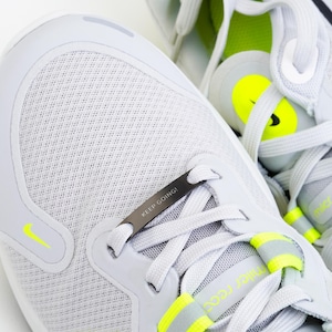 Puede incluir: Un par de zapatillas de running blancas con un acento negro y amarillo. Las zapatillas tienen un cierre de cordones de metal con las palabras "KEEP GOING!" grabadas en &eacute;l.