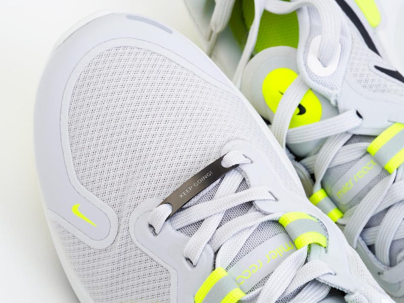 Puede incluir: Un par de zapatillas de running blancas con un acento negro y amarillo. Las zapatillas tienen un cierre de cordones de metal con las palabras "KEEP GOING!" grabadas en &eacute;l.