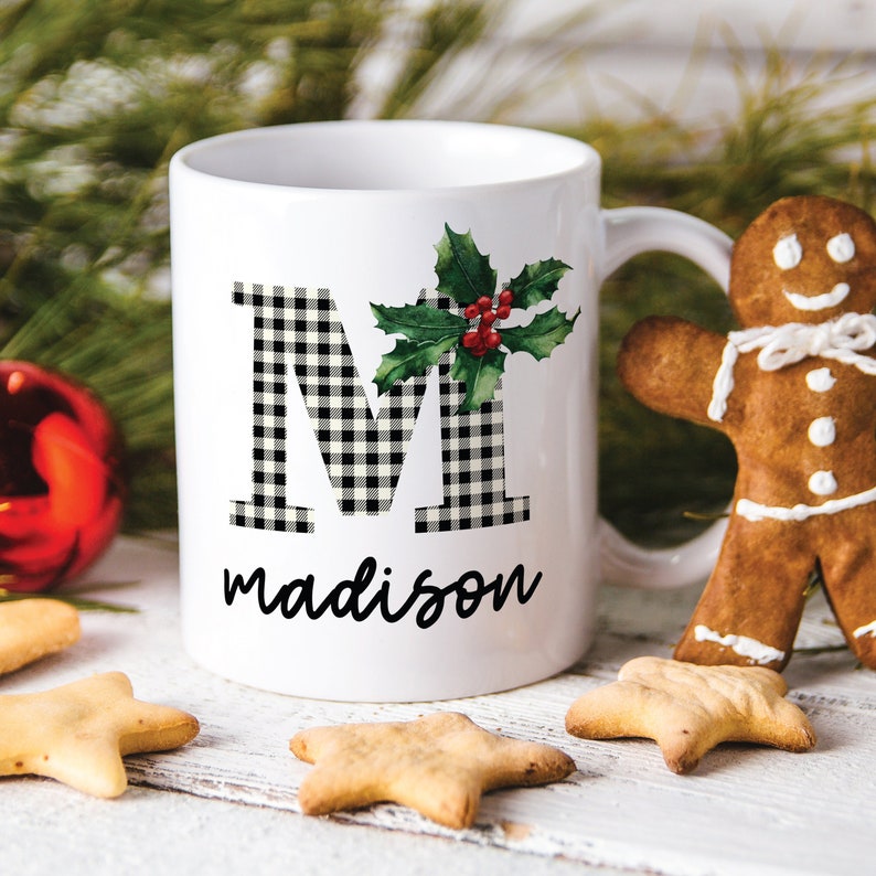 Puede incluir: Taza de cer&aacute;mica blanca con una letra M a cuadros en blanco y negro y la palabra "madison" impresa debajo. Una rama de acebo verde con bayas rojas est&aacute; a la derecha de la letra M. La taza tiene un asa negra.