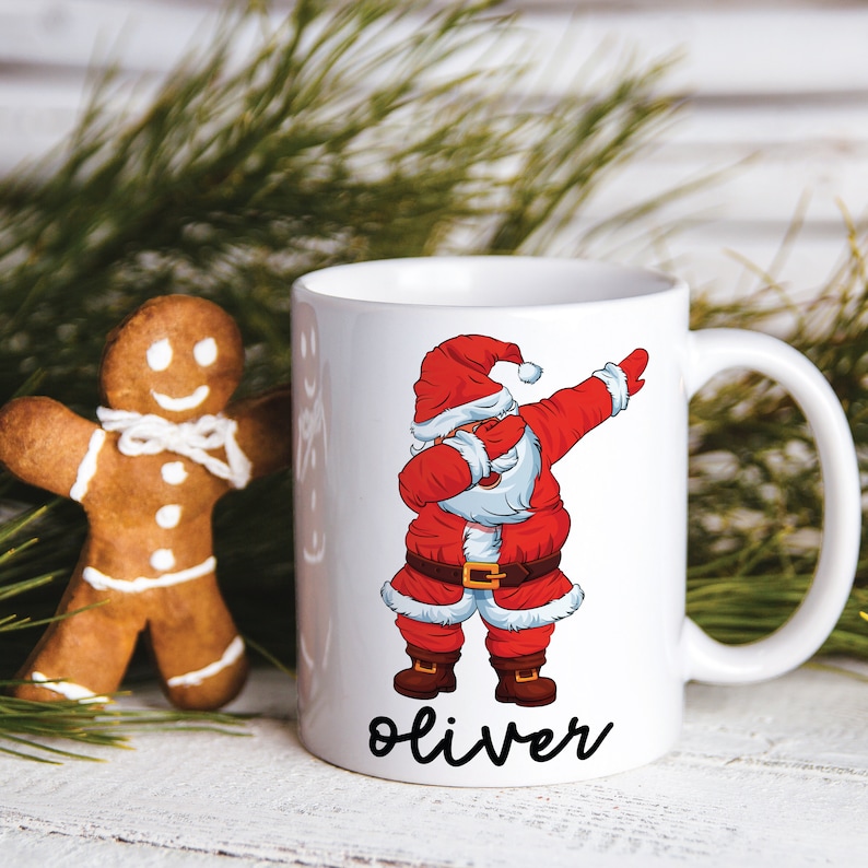 Puede incluir: Taza de cer&aacute;mica blanca con una ilustraci&oacute;n de Pap&aacute; Noel rojo y blanco haciendo el dab. Pap&aacute; Noel lleva un traje rojo con ribete blanco y un cintur&oacute;n negro. La taza tiene el nombre "Oliver" escrito en cursiva negra en la parte inferior.
