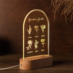 May include: A personalized LED night light with a wooden base. The clear acrylic panel features the text "Grandma's Garden" and floral illustrations with names. The light emits a warm glow, perfect for a bedside table.