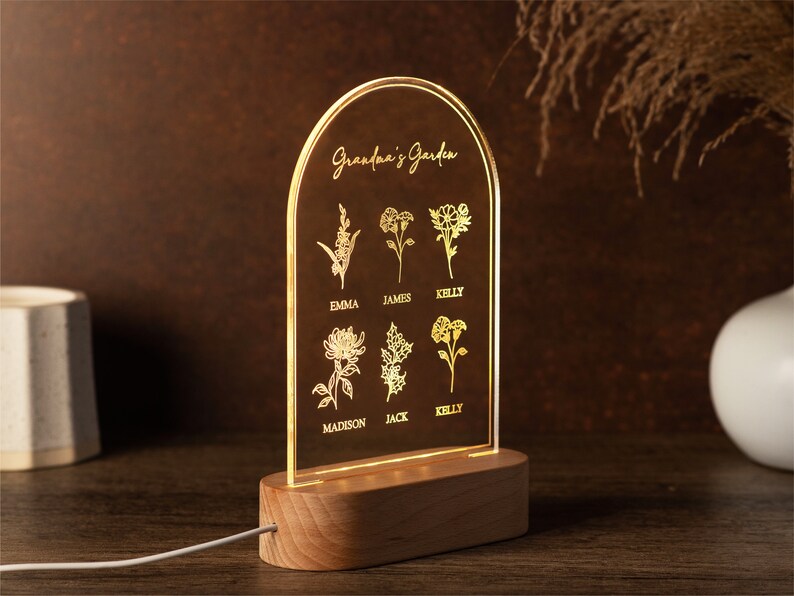 May include: A personalized LED night light with a wooden base. The clear acrylic panel features the text "Grandma's Garden" and floral illustrations with names. The light emits a warm glow, perfect for a bedside table.