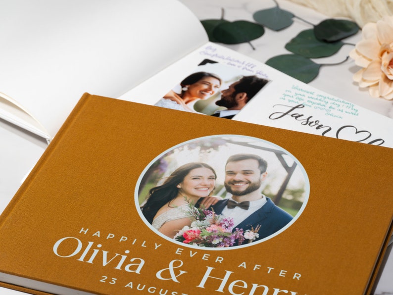 May include: A brown photo album with a circular cutout on the front cover. The cutout shows a photo of a couple smiling at the camera. The text "Happily Ever After Olivia & Henry 23 August" is printed on the front cover.