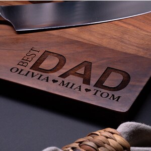 May include: A wooden cutting board with the text "Best Dad" engraved on it. The names Olivia, Mia, and Tom are also engraved on the board, along with hearts.