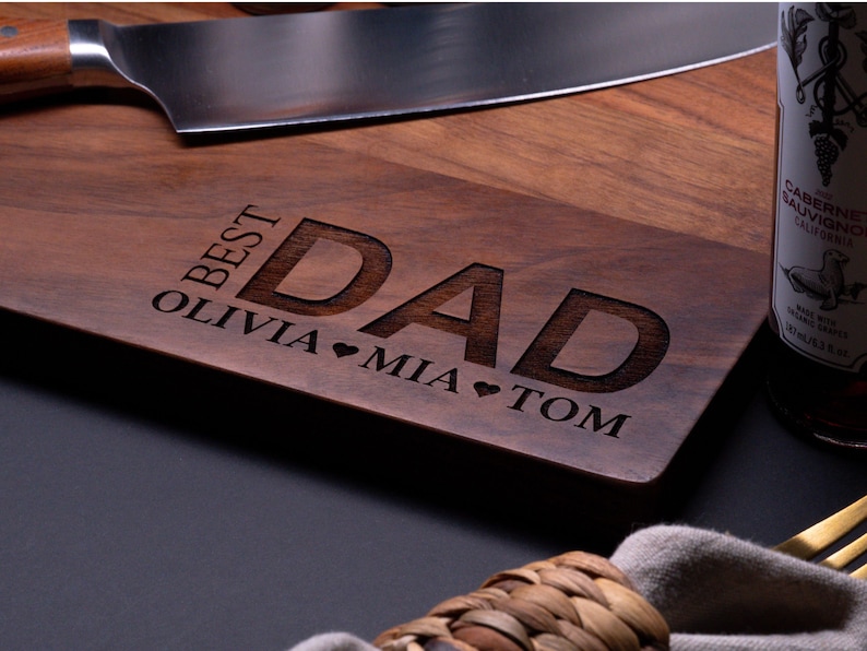 May include: A wooden cutting board with the text "Best Dad" engraved on it. The names Olivia, Mia, and Tom are also engraved on the board, along with hearts.