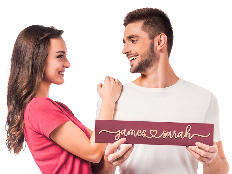 May include: A couple holding a burgundy sign with the text "james &hearts; sarah" in gold lettering.