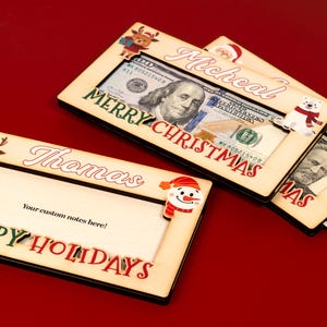 May include: Three wooden picture frames with a Christmas theme. Each frame has a different holiday character and the words "Merry Christmas" or "Happy Holidays". The frames are designed to hold cash or gift cards.