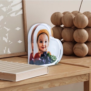 K&ouml;nnte beinhalten: Ein transparenter herzf&ouml;rmiger Bilderrahmen mit einem Foto eines jungen M&auml;dchens, das einen Strohhut tr&auml;gt. Der Rahmen steht auf einem Holztisch.