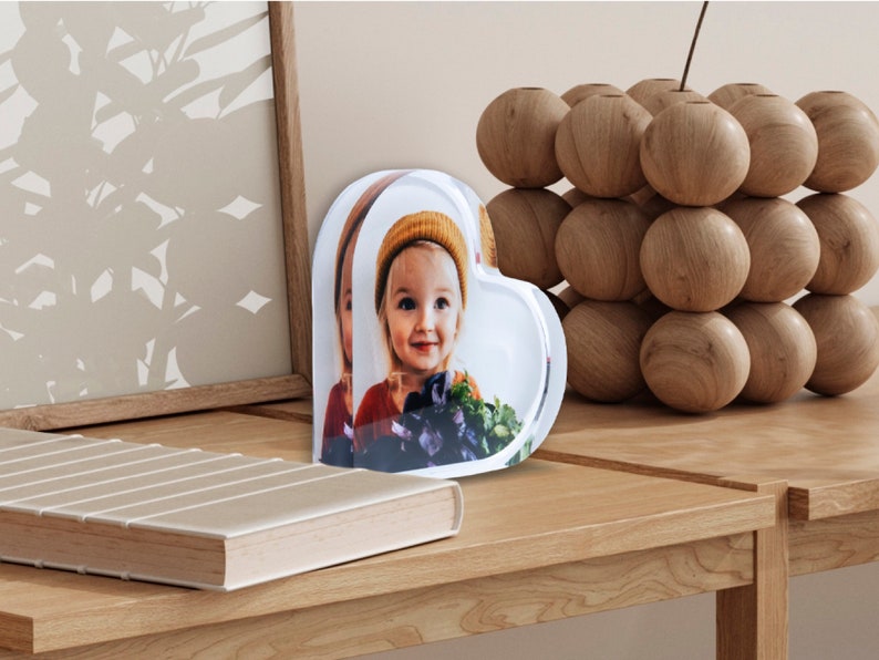 K&ouml;nnte beinhalten: Ein transparenter herzf&ouml;rmiger Bilderrahmen mit einem Foto eines jungen M&auml;dchens, das einen Strohhut tr&auml;gt. Der Rahmen steht auf einem Holztisch.