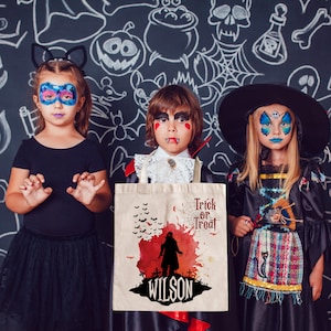 May include: Three children dressed in Halloween costumes, a girl in a black cat costume, a boy in a vampire costume, and a girl in a witch costume. The boy is holding a white canvas tote bag with a red and black Halloween design and the text "Trick or Treat" and "Wilson".