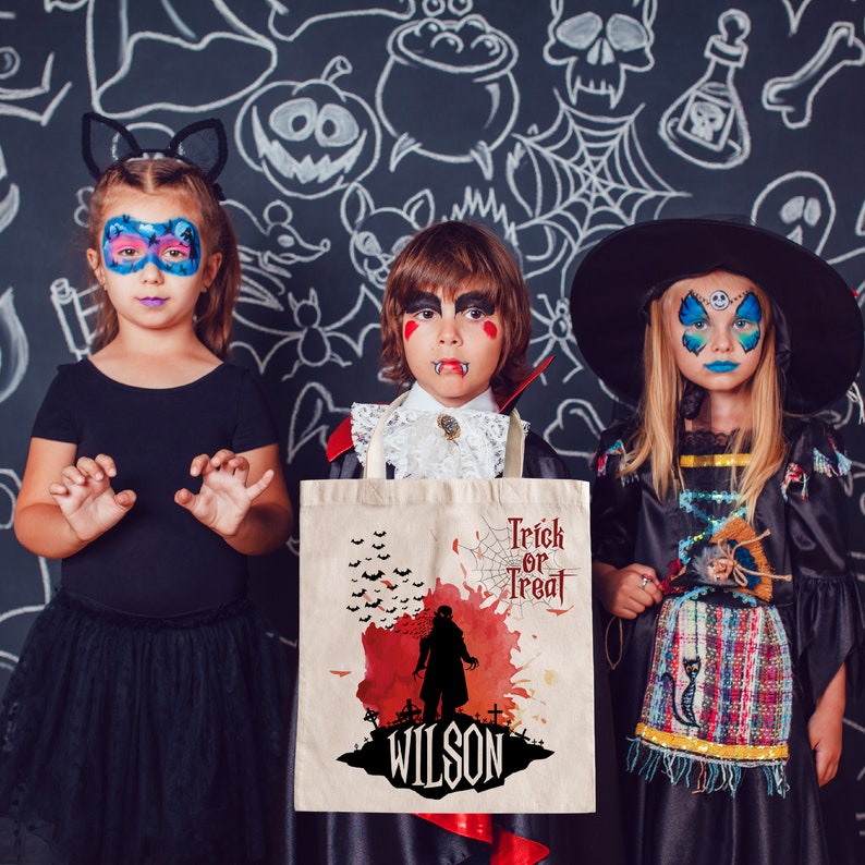May include: Three children dressed in Halloween costumes, a girl in a black cat costume, a boy in a vampire costume, and a girl in a witch costume. The boy is holding a white canvas tote bag with a red and black Halloween design and the text "Trick or Treat" and "Wilson".