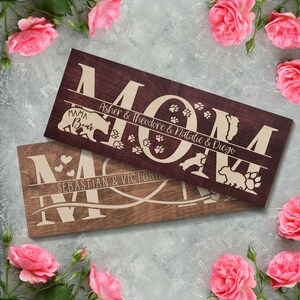 May include: Two wooden signs with the word "MOM" in large letters. The top sign is stained a dark brown and features the names "Asher & Theodore & Natalie & Diego" and paw prints. The bottom sign is a lighter brown and features the names "Sebastian & Victoria" and hearts.