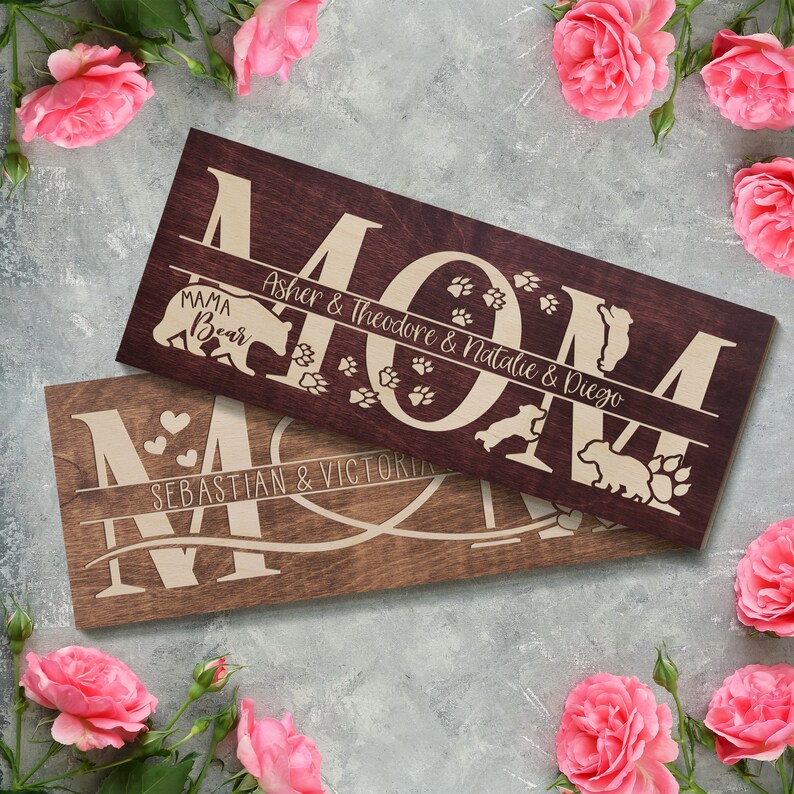 May include: Two wooden signs with the word "MOM" in large letters. The top sign is stained a dark brown and features the names "Asher & Theodore & Natalie & Diego" and paw prints. The bottom sign is a lighter brown and features the names "Sebastian & Victoria" and hearts.