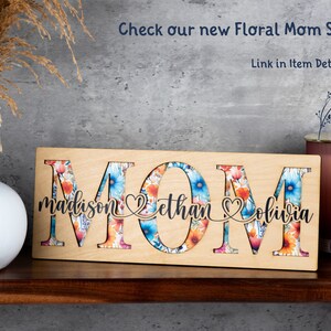 May include: Wooden sign with the word "MOM" in large letters. The letters are cut out and filled with a floral pattern. The names "madison", "ethan", and "olivia" are written below the letters.