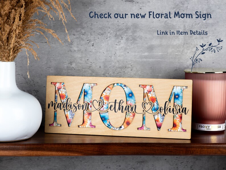 May include: Wooden sign with the word "MOM" in large letters. The letters are cut out and filled with a floral pattern. The names "madison", "ethan", and "olivia" are written below the letters.